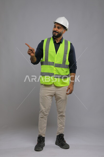 التقدم والتطور في المجال الهندسي السعودي، بورتريه لمهندس عربي سعودي خليجي يرتدي الخوذة وسترة الحماية  يشير بإصبعه الى مكان ما، أهمية الهندسة في تطوير وتحسين البنية التحتية للمدن، صورة الجسم بالطول الكامل، خلفية رمادية