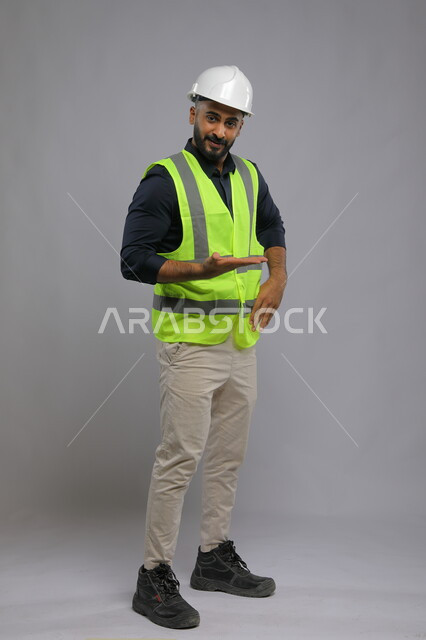 التدقيق والإشراف على العمل، بورتريه لمهندس عربي سعودي خليجي يرتدي الخوذة وسترة الحماية وراحة يده مفتوحة للأعلى، أهمية الهندسة في تحسين حياة البشر، صورة الجسم بالطول الكامل، خلفية رمادية