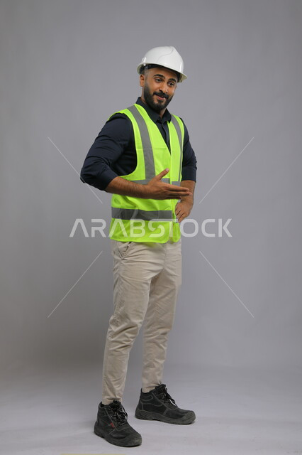  التقدم والتطور في المجال الهندسي السعودي، بورتريه لمهندس عربي سعودي خليجي يرتدي الخوذة وسترة الحماية يشير بيده إلى مكان ما، تصميم وتنفيذ المشاريع الهندسية الكبرى، صورة الجسم بالطول الكامل، خلفية رمادية