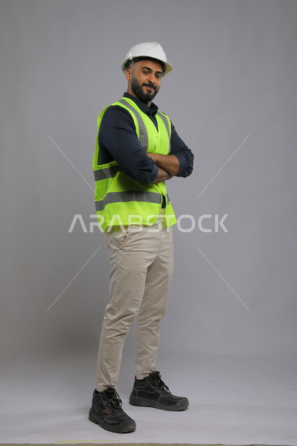 تعابير وجه تدل على الثقة بالنفس، التقدم والتطور في المجال الهندسي السعودي، بورتريه لمهندس عربي سعودي خليجي يرتدي الخوذة وسترة الحماية متكتف اليدين ينظر الى الكاميرا بابتسامة، تصميم وتنفيذ المشاريع الهندسية الكبرى، صورة الجسم بالطول الكامل، خلفية رمادية