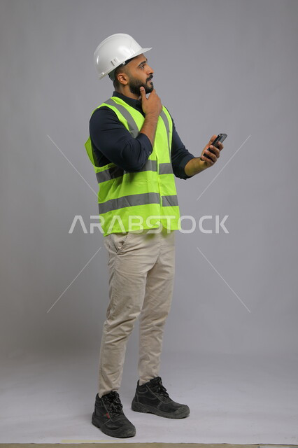 ايماءات التأمل والتفكير، دمج التكنولوجيا والتقنية مع البيئة العملية، متابعة المشاريع وتدقيق العمل، بورتريه لمهندس عربي سعودي خليجي يرتدي الخوذة وسترة الحماية ويمسك بيده الهاتف المحمول،  صورة الجسم بالطول الكامل، خلفية رمادي