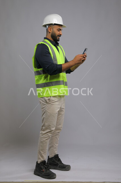 دور التكنولوجيا في تطوير الهندسة وتحسين الإنتاجية، بورتريه لمهندس عربي سعودي خليجي يرتدي الخوذة وسترة الحماية ويقوم باستخدام الهاتف المحمول، دور الهندسة في تقدم المجتمع السعودي، صورة الجسم بالطول الكامل، خلفية رمادي