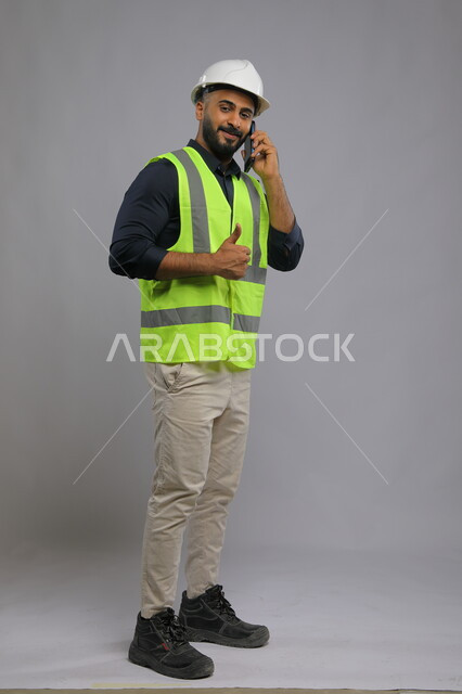 ايماءات القبول والموافقة، الاشراف والرقابة على المشاريع الهندسية، بورتريه لمهندس عربي سعودي خليجي يرتدي الخوذة وسترة الحماية ويصنع بيده اشارة الاعجاب، دور الهندسة في تقدم المجتمع السعودي، صورة الجسم بالطول الكامل، خلفية رمادي