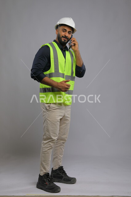 Field of construction and engineering projects, supervision and control of engineering projects, portrait of a Saudi Arabian Gulf engineer wearing a helmet and protective vest and talking on a mobile phone, full-length body portrait, gray background