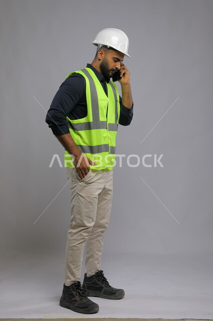 الاشراف والرقابة على المشاريع الهندسية، بورتريه لمهندس عربي سعودي خليجي يرتدي الخوذة وسترة الحماية ويتحدث على الهاتف المحمول، مجال الانشاءات والمشاريع الهندسية, صورة الجسم بالطول الكامل، خلفية رمادي