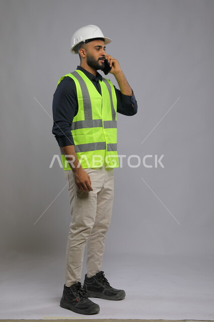 Supervision and control of engineering projects, portrait of a Saudi Arabian Gulf engineer wearing a helmet and protection vest and talking on a mobile phone, field of construction and engineering projects, full-length body portrait, gray background