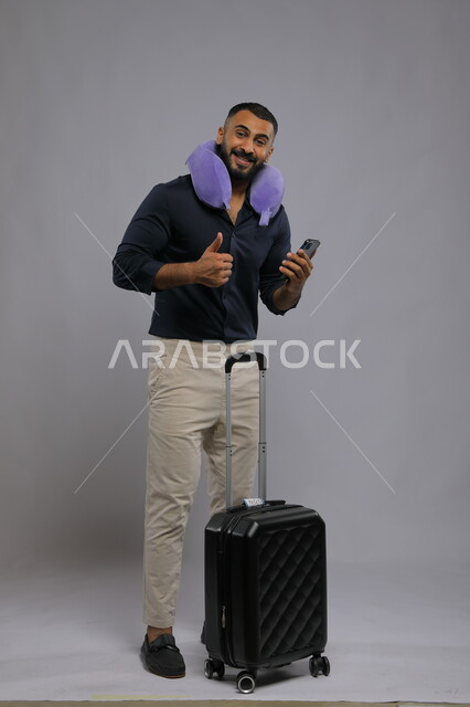 ايماءات الاعجاب والقبول، استخدام تطبيقات الهاتف لشراء تذكرة الطيران، الإستعداد للرحلة السياحية، بورتريه لرجل عربي سعودي خليجي يرتدي ملابس كاجوال يضع وسادة النوم و يمسك الهاتف المحمول بجانبه حقيبة السفر، صورة الجسم بالطول الكامل، خلفية رمادية