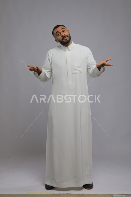 الشعور بالحيرة وعدم المعرفة، بورتريه لرجل عربي سعودي خليجيي يرتدي الثوب التقليدي يرفع يفتح كفيه بإيماءات عدم التأكد, التردد وعدم الثقة بخصوص أمر معين، الوقوف باستقامة والنظر إلى الكاميرا، صورة الجسم بالطول الكامل,خلفية رمادية