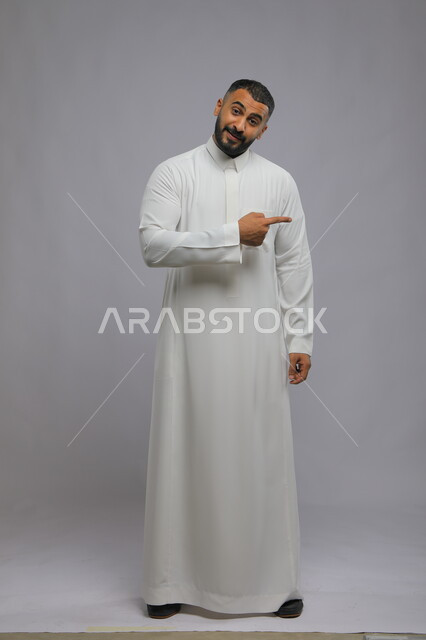 إيماءات الرضا والسرور، بورتريه لرجل عربي خليجي سعودي مبتسم يرتدي الزي التقليدي  يشير بإصبعه نحو شئ محدد، الاشارة تجاه هدف معين أو شخص ما،الوقوف باستقامة والنظر إلى الكاميرا،  صورة الجسم بالطول الكامل، خلفية رمادية.