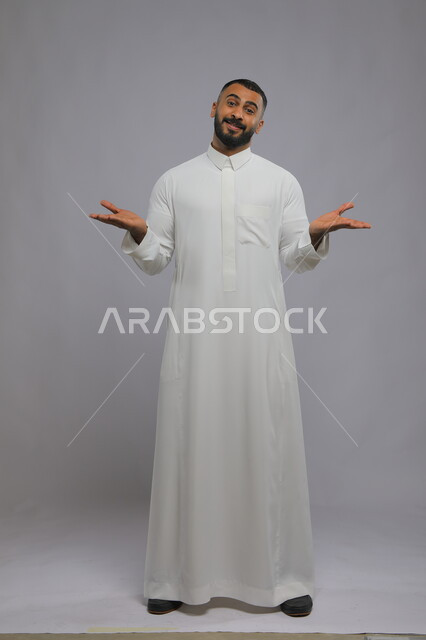 Hesitation and lack of confidence regarding a certain matter, portrait of a Saudi Gulf Arab man wearing a traditional dress, raising and opening his palms with gestures of uncertainty, feeling of confusion and lack of knowledge, standing straight and looking at the camera, full-length body portrait, gray background