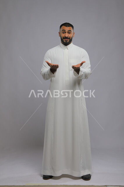 إيماءات التأكيد وتحديد الشيء المقابل, بورتريه لرجل عربي سعودي خليجيي يرتدي الثوب التقليدي يرفع يديه للإشارة إلى شيء ما, الثقة في معرفة أمر معين, صورة الجسم بالطول الكامل,خلفية رمادية