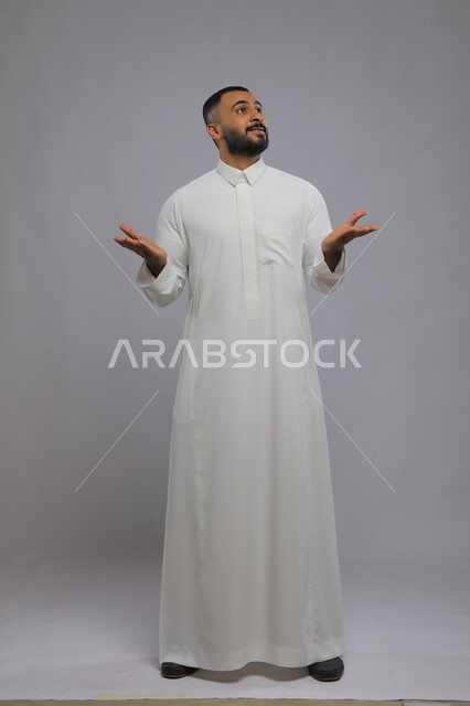 الشكر والامتنان على المعروف ، التساؤل عن حدوث أمر ما، بورتريه لشاب عربي سعودي خليجي يرتدي الثوب التقليدي يرفع كفيه وينظر الى الأعلى ، انتظار شيء ما ،الإعجاب والمدح بشيء جميل, صورة الجسم بالطول الكامل, خلفية رمادية.