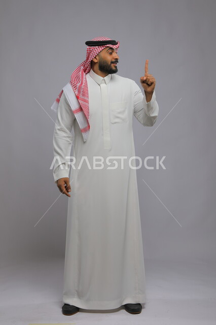 Expressions indicating warning and caution, standing up straight with inquiring gestures and pointing to something, portrait of a smiling Saudi Gulf Arab man wearing a shemagh and traditional thobe, raising his finger high, full-length body portrait, gray background