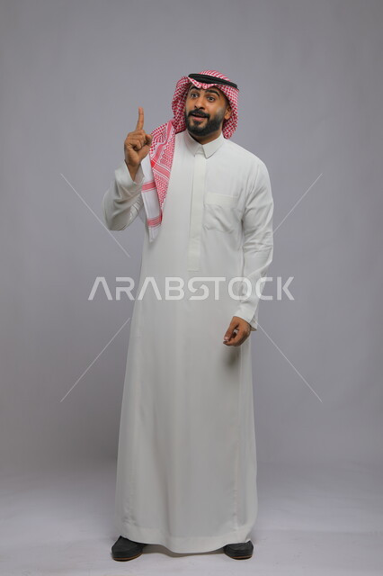 ايماءات الاستفسار والاشارة إلى شيء ما, الوقوف باستقامة والنظر إلى الأعلى, بورتريه لرجل عربي خليجي سعودي مبتسم يرتدي الشماغ والثوب التقليدي يرفع اصبعه عاليا بإيماءات التوضيح, صورة الجسم بالطول الكامل , خلفية رمادية
