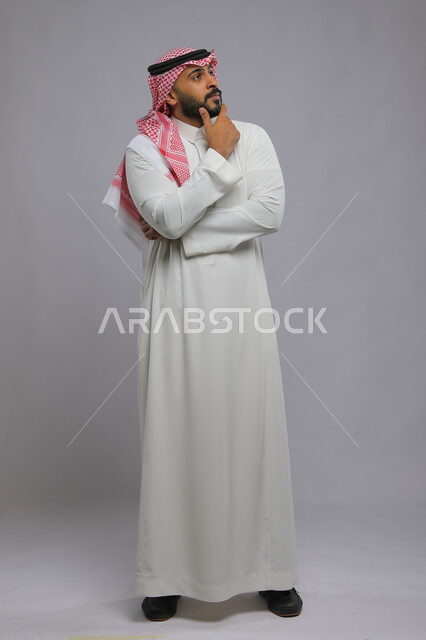 التأمل والتخطيط للمستقبل، إيجاد حل لمشكلة ما، بورتريه لشاب عربي خليجي سعودي يرتدي الثوب والشماغ التقليدي يضع يده على ذقنه و ينظر إلى شيء ما بإيماءات التفكير، التمعن والتركيز وشرود الذهن ، صورة الجسم بالطول الكامل، خلفية رمادية