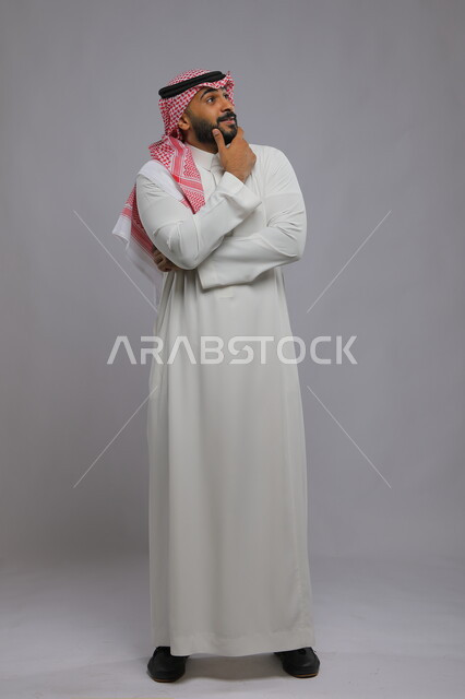  التمعن والتركيز وشرود الذهن ،التأمل والتخطيط للمستقبل، إيجاد حل لمشكلة ما، بورتريه لشاب عربي خليجي سعودي مبتسم يرتدي الثوب والشماغ التقليدي يضع يده على ذقنه و ينظر إلى شيء ما بإيماءات التفكير، صورة الجسم بالطول الكامل، خلفية رمادية