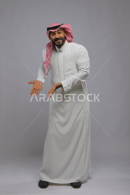  حركات تدل على هذا المكان (هنا)، تحديد موقع معين بإيماءات السرور والبشاشة ،بورتريه لشاب عربي خليجي سعودي مبتسم يرتدي الشماغ والثوب التقليدي يشير بيديه نحو الأسفل وينظر إلى الكاميرا، صورة الجسم بالطول الكامل، خلفية رمادية