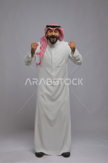 الاندماج بتشجيع فريق كرة القدم، حركات تدل على الكسب والربح وانجاز المهام ،ايماءات الفوز والانتصار ،بورتريه لشاب عربي خليجي سعودي يرتدي الشماغ والزي التقليدي يرفع يديه بتفاعل وحماس ،صورة الجسم بالطول الكامل، خلفية رمادية