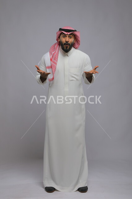 التفاجؤ عند فعل شيء مهم و النظر إلى الكاميرا ,الشعور بالصدمة و الدهشة والتعجب والذهول , بورتريه لشاب عربي خليجي سعودي يرتدي الشماغ والزي التقليدي و يرفع يديه للأمام بإيماءات الفرح والسرور ،صورة الجسم بالكامل, خلفية رمادية