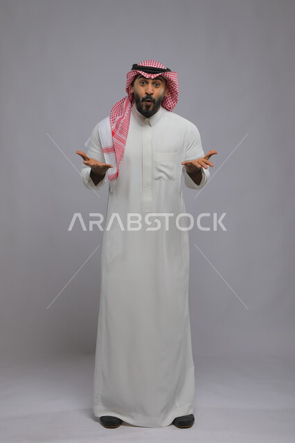 Looking at the camera with gestures of joy and pleasure, feeling shock, astonishment, astonishment and astonishment, portrait of a Saudi Gulf Arab young man wearing a shemagh and traditional dress and raising his hands forward with an expression of surprise when doing something important, full body portrait, gray background