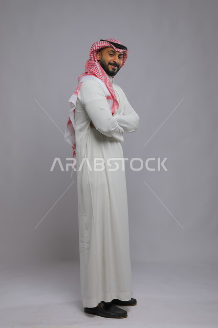 مفهوم الاهتمام بالمظهر الخارجي والشياكة ،شخصيه سعوديه مهمة ،النظر إلى الكاميرا بإيماءات أيدي متقاطعه والثقه والاعتزاز بالنفس، بورتريه من الجانب لرجل عربي خليجي سعودي مبتسم يرتدي الثوب والشماغ التقليدي ،صورة الجسم بالكامل، خلفية رمادية