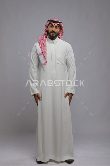 Self-confidence and a sense of strength, the concept of masculinity and elegance, standing upright and disciplined, paying attention to external appearance, a portrait of a smiling Saudi Arabian Gulf gentleman wearing a shemagh and a traditional thobe, placing his hands at his side and looking at the camera with gestures of pleasure, full-length body portrait, gray background.
