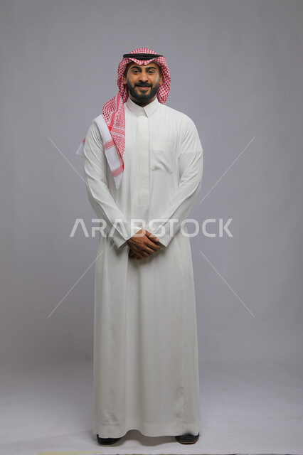 Standing upright with coherent hand movements, the concept of masculinity and elegance, attention to external appearance, a portrait of a smiling Saudi Gulf Arab man wearing traditional clothing looking at the camera with gestures of pleasure, full-length body portrait, gray background.