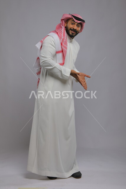 تحديد موقع معين بإيماءات السرور والبشاشة ، حركات تدل على هذا المكان (هنا)، بورتريه لشاب عربي خليجي سعودي مبتسم يرتدي الشماغ والثوب التقليدي يشير بيديه نحو الأسفل وينظر إلى الكاميرا، صورة الجسم بالطول الكامل، خلفية رمادية