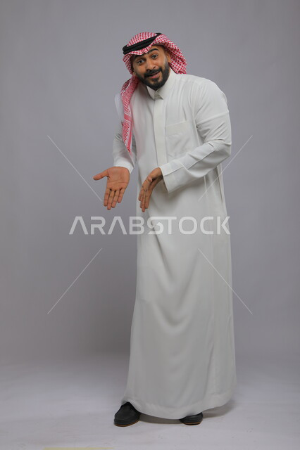 السرور والبشاشة وتحديد موقع معين، حركات تدل على هذا المكان (هنا)، بورتريه لشاب عربي خليجي سعودي مبتسم يرتدي الشماغ والثوب التقليدي يشير بيديه نحو الأسفل وينظر إلى الكاميرا، صورة الجسم بالطول الكامل، خلفية رمادية