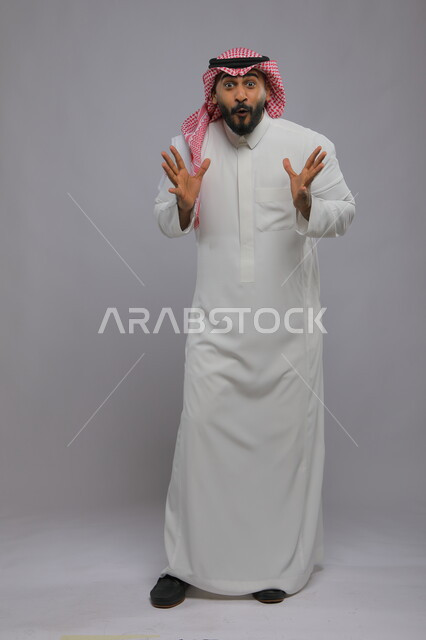 الشعور بالصدمة و الدهشة والتعجب والذهول , بورتريه لشاب عربي خليجي سعودي يرتدي الشماغ والزي التقليدي و يرفع يديه للأعلى بتعابير التفاجؤ عند رؤية شيء ما, النظر إلى الكاميرا بإيماءات الفرح والسرور , صورة الجسم بالكامل, خلفية رمادية