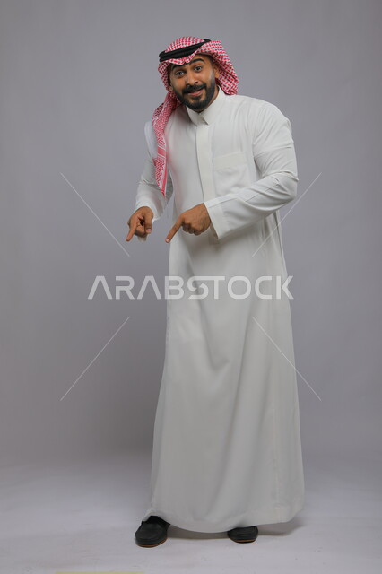 حركات تدل على هذا المكان (هنا)، البشاشة والسرور وتحديد موقع معين، بورتريه لشاب عربي خليجي سعودي مبتسم يرتدي الشماغ والثوب التقليدي يشير بأصابعه نحو الأسفل وينظر إلى الكاميرا، صورة الجسم بالطول الكامل، خلفية رمادية