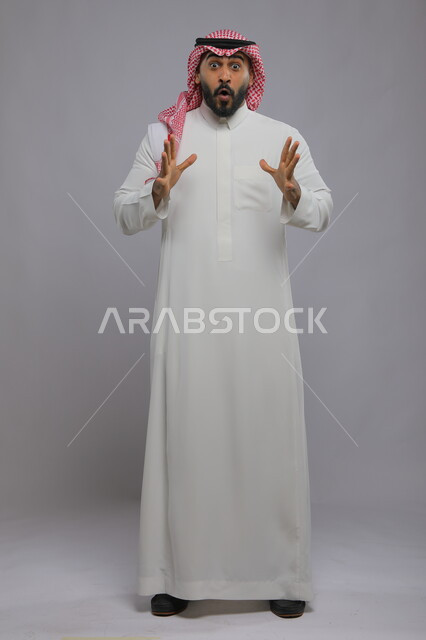 الشعور بالدهشة والتعجب والصدمة والذهول , التفاجؤ من شيء ما، بورتريه لشاب عربي خليجي سعودي يرتدي الشماغ والزي التقليدي و يرفع يديه للأعلى بتعابير النجاح وتحقيق الأهداف, النظر إلى الكاميرا بإيماءات الفرح والسرور , صورة الجسم بالكامل, خلفية رمادية