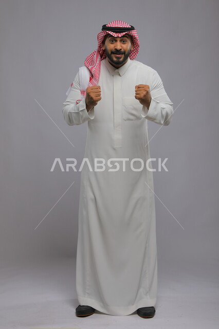حركات تدل على الربح وانجاز المهام والكسب ،ايماءات الفوز والانتصار ،بورتريه لشاب عربي خليجي سعودي يرتدي الشماغ والزي التقليدي يرفع يديه بتفاعل وحماس ،الاندماج بتشجيع فريق كرة القدم، صورة الجسم بالطول الكامل، خلفية رمادية
