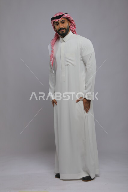 تعابير الفرح بالحصول على مايفتش عنه ، ايماءات البحث عن شيء ما في جيوب الثوب، بورتريه لشاب عربي خليجي سعودي مبتسم يرتدي الشماغ والزي التقليدي ينظر إلى الكاميرا، مفهوم الأناقة والاهتمام بالمظهر الخارجي، صورة الجسم بالطول الكامل، خلفية رمادية