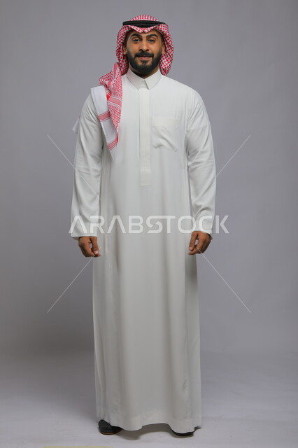 The concept of masculinity and elegance, attention to external appearance, a portrait of a smiling Saudi Gulf Arab young man wearing a shemagh and traditional dress, standing upright and looking at the camera with pleasure gestures, full-length body portrait, gray background.