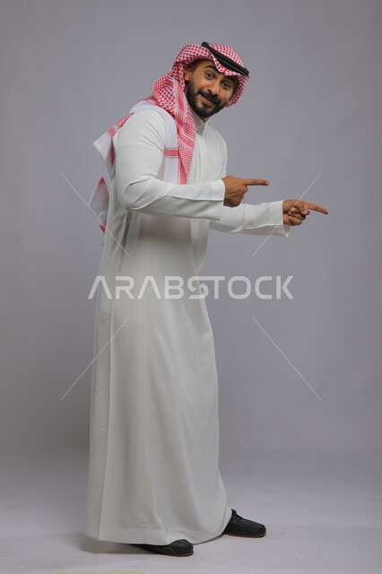 حركات تدل على الذهاب إلى مكان ما،  السعاده و السرور وتحديد موقع معين، بورتريه لشاب عربي خليجي سعودي مبتسم يرتدي الشماغ والثوب التقليدي يشير بأصابعه نحو الأسفل وينظر إلى الكاميرا، صورة الجسم بالطول الكامل، خلفية رمادية