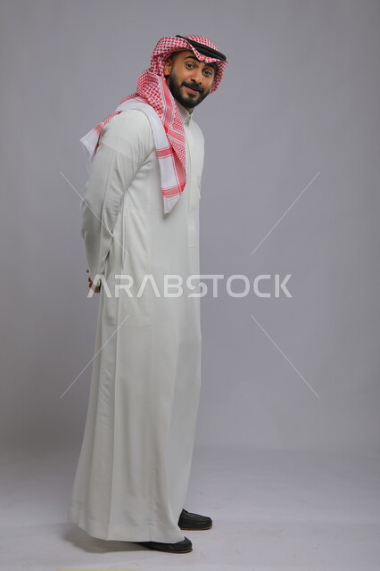  ايماءات السرور والسعادة، بورتريه لشاب عربي خليجي سعودي مبتسم يرتدي الشماغ والثوب التقليدي يضع يديه وراء ظهره وينظر الى الكاميرا، مفهوم الاناقة والاهتمام بالمظهر الخارجي، صورة الجسم بالطول الكامل، خلفية رمادية