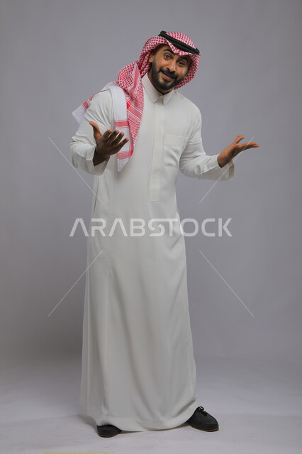 تعابير الاعجاب والمدح لشيء جميل، التساؤل والاستفهام عن سبب حدوث أمر معين، بورتريه لشاب عربي خليجي سعودي مبتسم يرتدي الشماغ والثوب التقليدي يقوم برفع الكفين والنظر الى الكاميرا بإيماءات السرور، صورة الجسم بالطول الكامل، خلفية رمادية