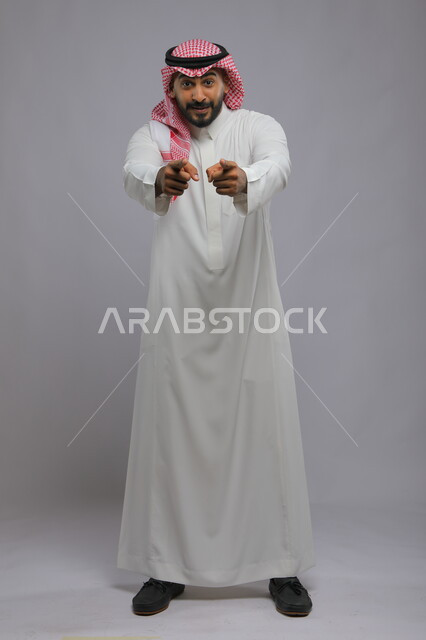 ايماءات التأكيد وتحديد الشيء المقابل, الإشارة الى شخص ما بحركات التواجد هنا, بورتريه لرجل عربي خليجي سعودي يرتدي الثوب التقليدي والشماغ يرفع اصابع السبابة للأمام بتعابير السرور, صورة الجسم بالطول الكامل , خلفية رمادية