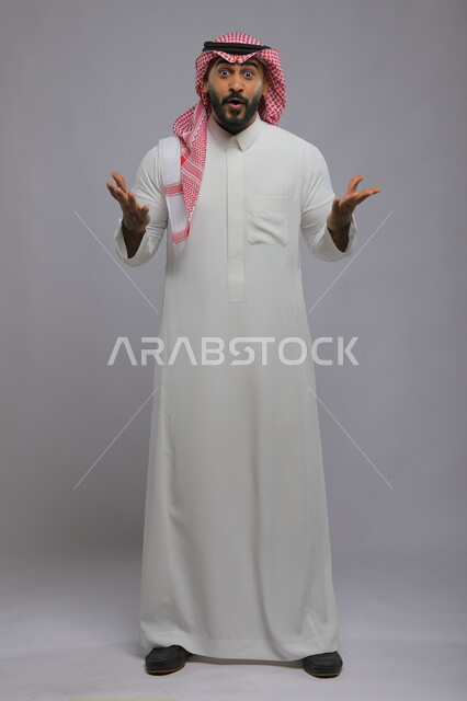 النظر إلى الكاميرا بإيماءات الفرح والسرور ,الشعور بالصدمة و الدهشة والتعجب والذهول , بورتريه لشاب عربي خليجي سعودي يرتدي الشماغ والزي التقليدي و يرفع يديه للأعلى بتعابير التفاجؤ عند رؤية شيء ما, صورة الجسم بالكامل, خلفية رمادية