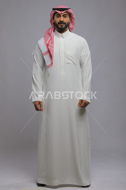 Concern with external appearance, the concept of masculinity and elegance, a portrait of a smiling Saudi Gulf Arab young man wearing a shemagh and traditional dress, standing upright and looking at the camera with pleasure gestures, full-length body portrait, gray background.