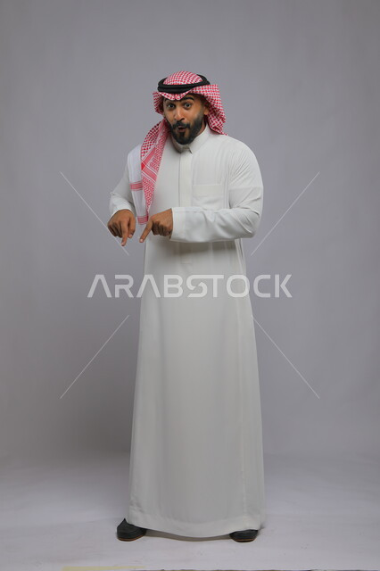 ايماءات الدهشة والذهول بوجود شيء ما، اشارات تدل على هذا المكان (هنا)، بورتريه لرجل عربي خليجي سعودي مبتسم يرتدي الشماغ والثوب التقليدي يشير بأصابعه نحو الأسفل ،النظر إلى الكاميرا و تحديد موقع معين، صورة الجسم بالطول الكامل، خلفية رمادية