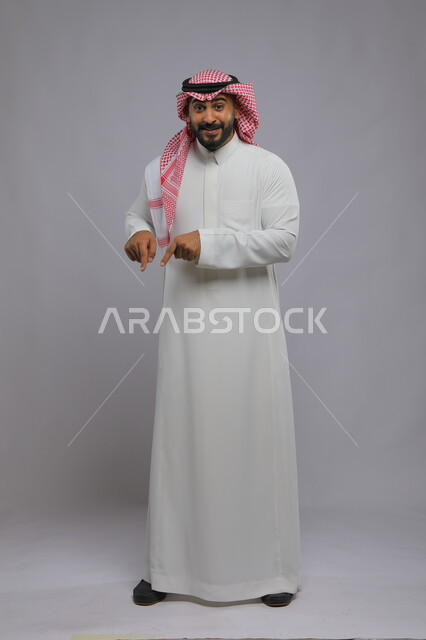 حركات تدل على وجود شيء ما، اشارات تدل على هذا المكان (هنا)، البشاشة والسرور وتحديد موقع معين، بورتريه لرجل عربي خليجي سعودي مبتسم يرتدي الشماغ والثوب التقليدي يشير بأصابعه نحو الأسفل وينظر إلى الكاميرا، صورة الجسم بالطول الكامل، خلفية رمادية