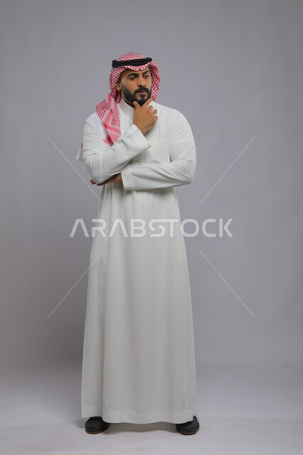 إيجاد حل لمشكلة ما، التركيز وشرود الذهن والتمعن ،التأمل والتخطيط للمستقبل، بورتريه لرجل عربي خليجي سعودي يرتدي الثوب والشماغ التقليدي يضع يده على ذقنه و ينظر إلى شيء ما بإيماءات التفكير، صورة الجسم بالطول الكامل، خلفية رمادية