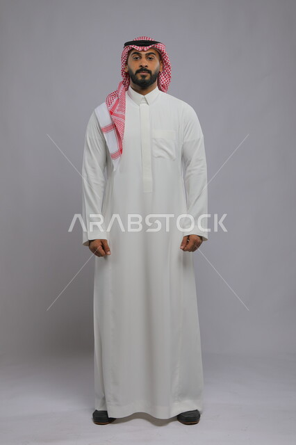 تعابير الجدية والثقة بالنفس، مفهوم الاهتمام بالمظهر الخارجي و الرجولة والأناقه، بورتريه لرجل عربي خليجي سعودي يرتدي الشماغ والثوب التقليدي يقف باستقامة والنظر إلى الكاميرا يإيماءات العبوس، صورة الجسم بالطول الكامل، خلفية رمادية.