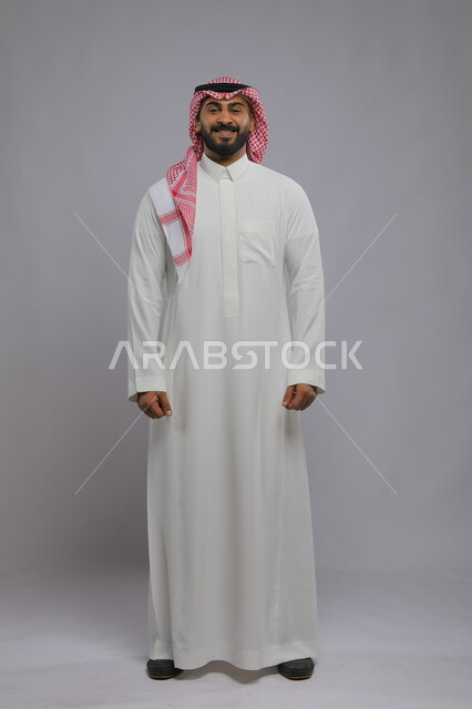 ايماءات البشاشة والضحك والسرور، مفهوم الاهتمام بالمظهر الخارجي و الرجولة والأناقه، بورتريه لشاب عربي خليجي سعودي مبتسم يرتدي الشماغ والثوب التقليدي يقف باستقامة وينظر إلى الكاميرا ، صورة الجسم بالطول الكامل، خلفية رمادية.