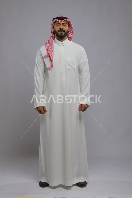 The concept of caring about external appearance, masculinity and elegance, a portrait of a smiling Saudi Gulf Arab young man wearing a shemagh and traditional dress, standing straight and looking at the camera with a gesture of pleasure, full-length body portrait, gray background.