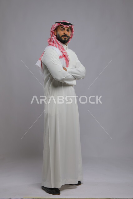 شخصية مهمة سعودية ،الوقوف بحركات أيدي متقاطعة، الاهتمام بالمظهر الخارجي، مفهوم الأناقة والرجولة، بورتريه لرجل عربي خليجي سعودي يرتدي الثوب والشماغ التقليدي ينظر إلى الكاميرا بإيماءات الثقه بالنفس، صورة لكامل الجسم، خلفية رمادية.