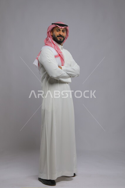 الوقوف بحركات أيدي متقاطعة، الاهتمام بالمظهر الخارجي، مفهوم الأناقة والرجولة، بورتريه لرجل عربي خليجي سعودي مبتسم يرتدي الثوب والشماغ التقليدي ينظر إلى الكاميرا بإيماءات السرور و الثقه بالنفس، صورة لكامل الجسم، خلفية رمادية.