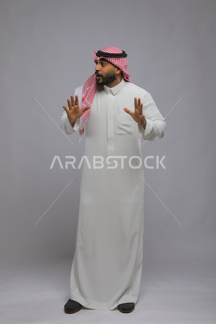 النظر إلى مكان معين بإيماءات الذعر والخوف ,الشعور بالصدمة و الدهشة والتعجب والذهول , بورتريه لشاب عربي خليجي سعودي يرتدي الشماغ والزي التقليدي و يرفع يديه للأعلى بتعابير التفاجؤ عند رؤية شيء ما, صورة الجسم بالكامل, خلفية رمادية